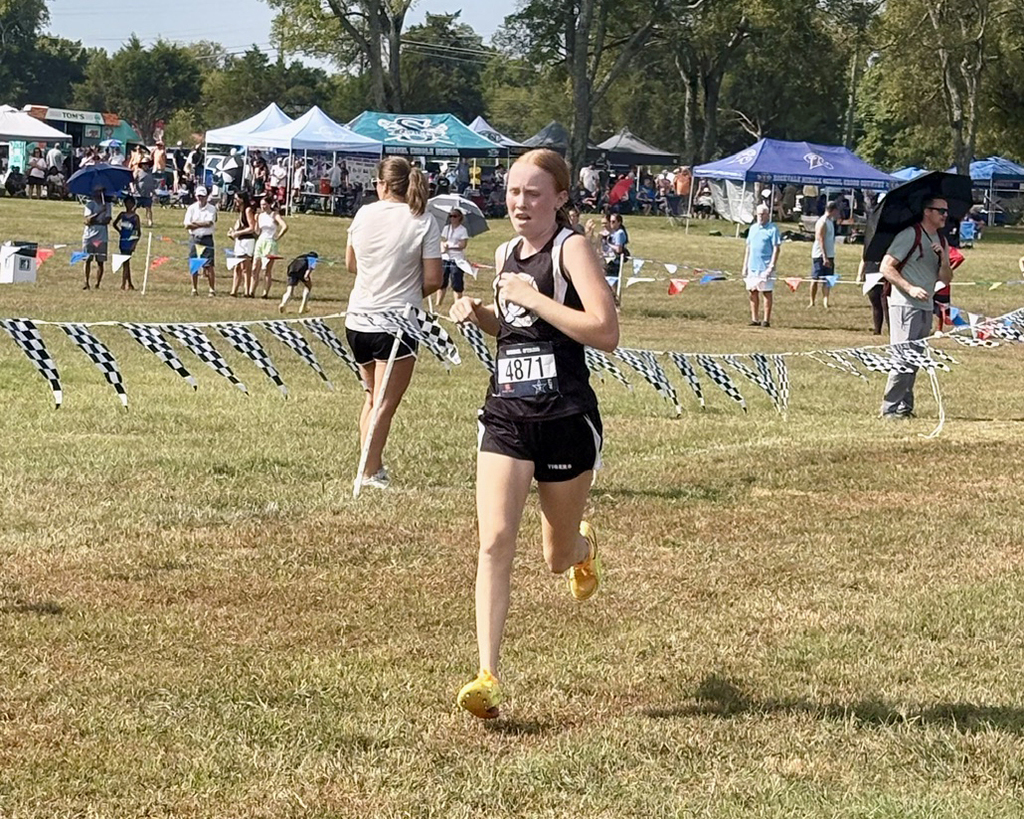 A runner in a black uniform with yellow shoes is sprinting in a grassy field during a race.