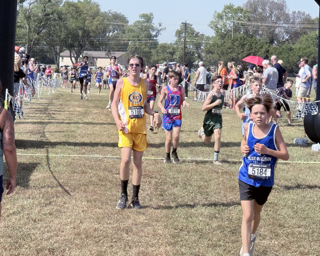 Runners in a cross-country event, including a man in yellow, compete on a field with a crowd watching.