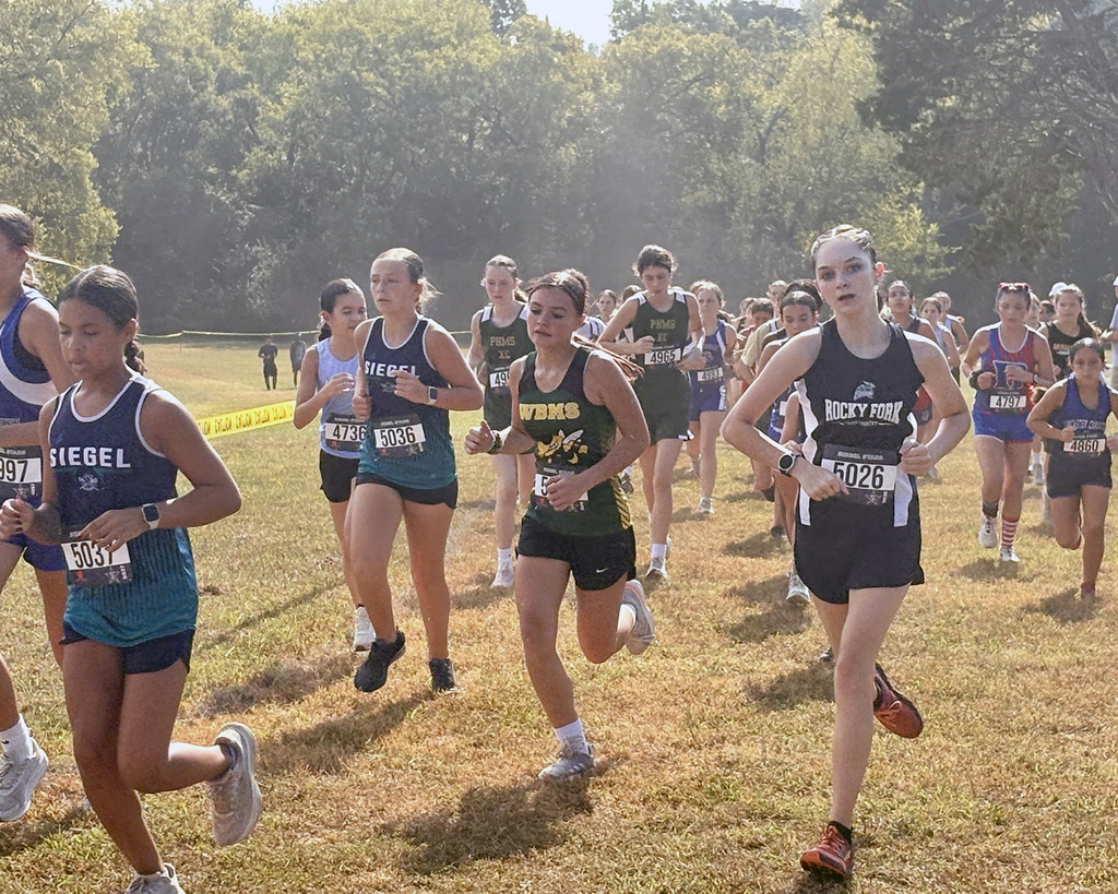 A group of athletes wearing uniforms runs in a race on a grassy field with trees in background.