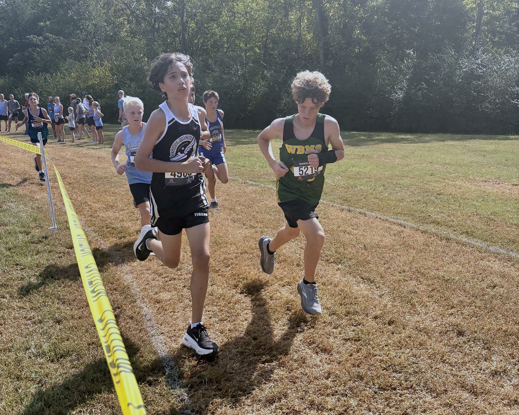Several children running in an outdoor cross-country race, with a yellow tape marking the route.