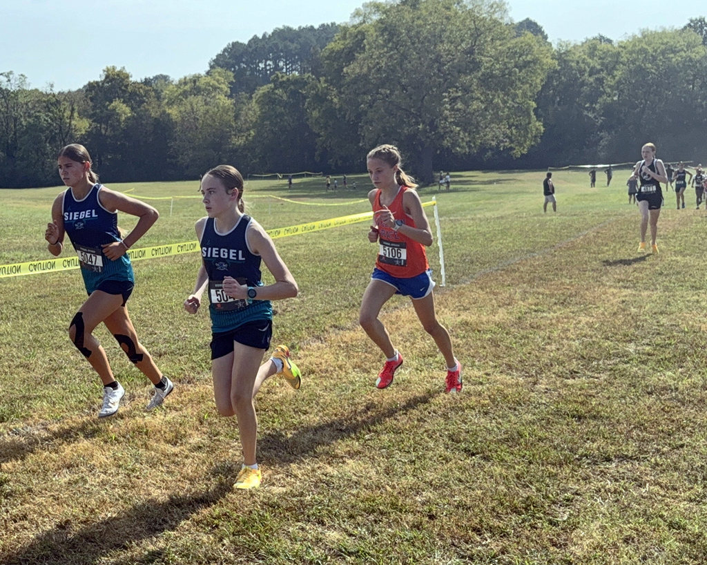 Three people in athletic wear run on a grassy field with trees in the background. They have numbered shirts and run near yellow caution tape.