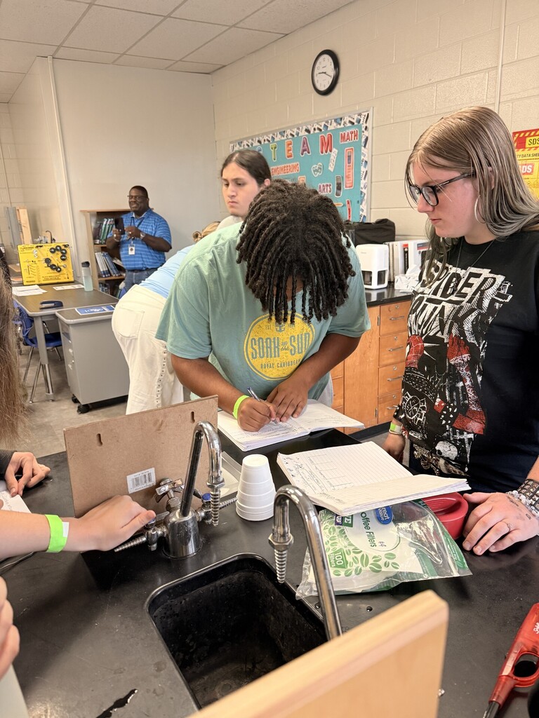 Four people in a lab. Two women are writing on paper. A man is writing on a box. A clock on the wall.