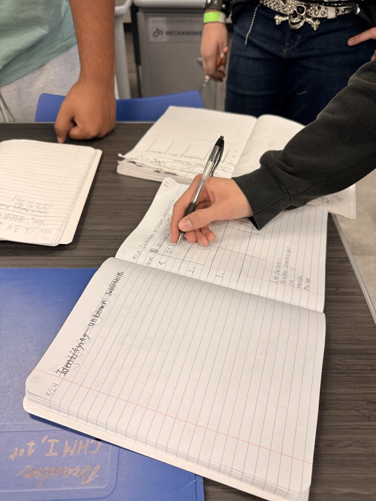 A person writes in a notebook. Several other notebooks and papers are on the table.