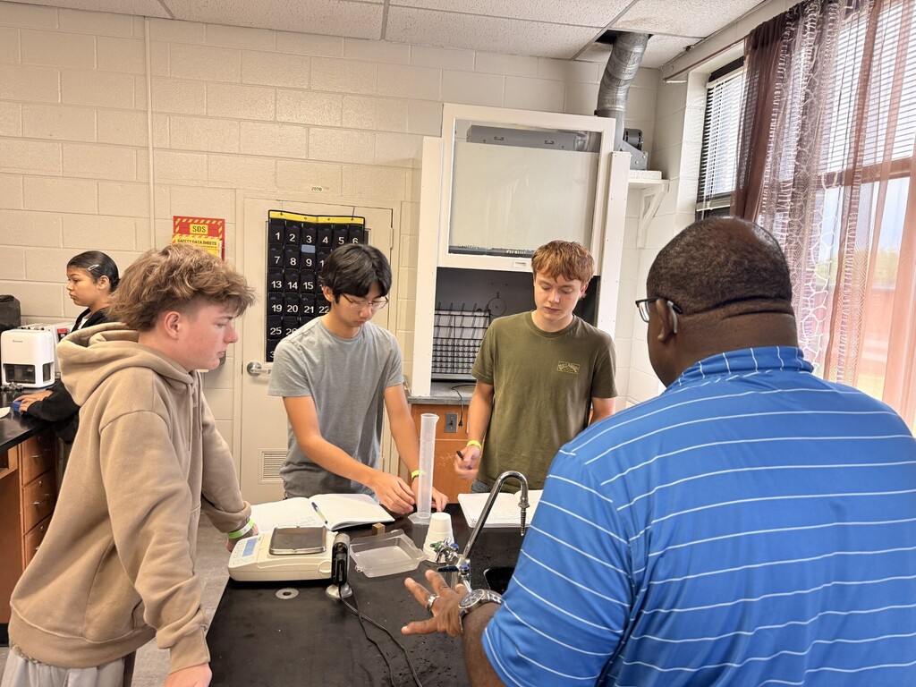 Five students in a science lab with a table, wires, and equipment. A teacher explains.