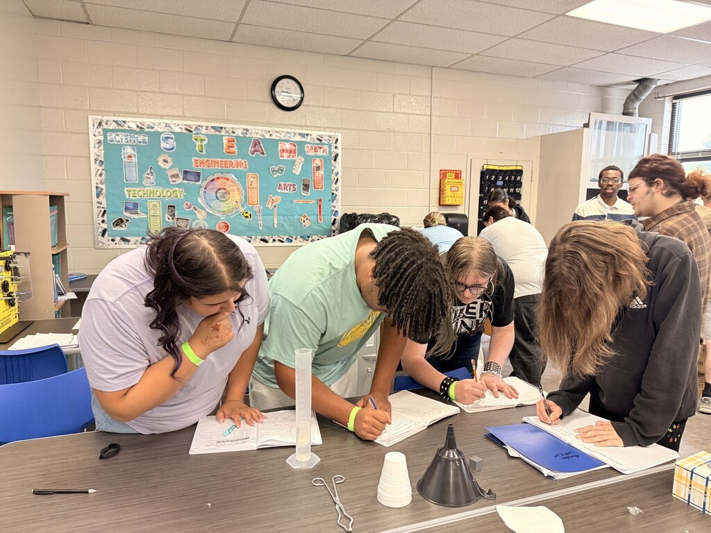 Group of students in a classroom bent over a table, writing on papers and handling objects.