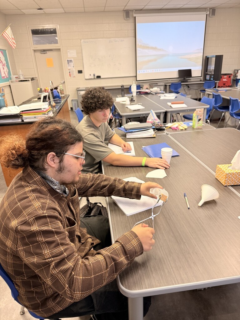 Two people sit at a table in a classroom, one with a ponytail and glasses, the other with a bracelet.
