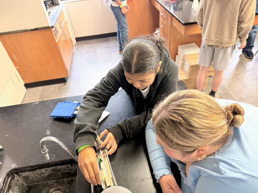 A person in a blue jacket and a person in a black jacket work at a sink in a lab.