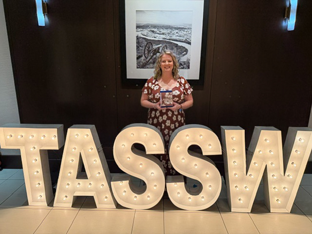 Delaina Kinnison holding her award behind large TASSW sign