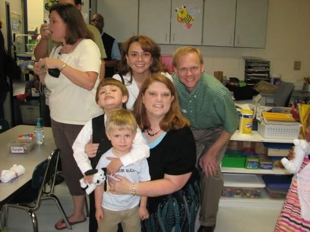 A family of four and two children stand together in a classroom, smiling and posing for a photo.