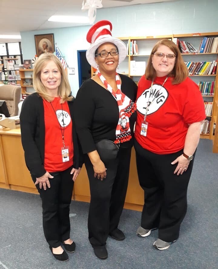 Three women wear red T-shirts and black pants. They stand in a room with bookshelves, a desk, and ceiling fan.