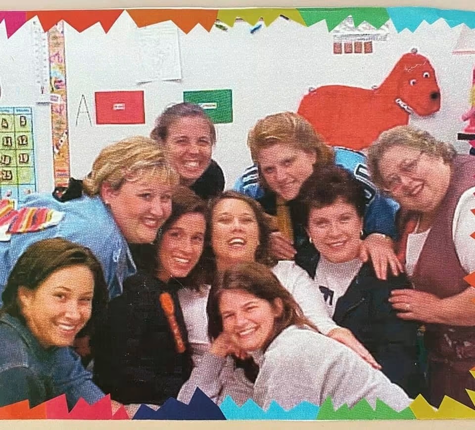 Group of women smiling for a photo in a classroom. Brightly colored decorations in background.