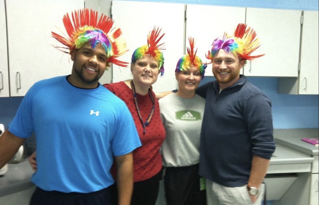 Four people in a kitchen wearing colorful headdresses. One man in blue, one in red, and two in white.