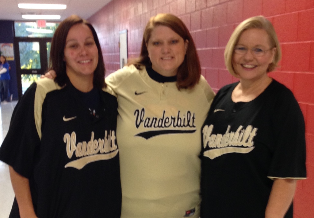 Three women in sports jerseys stand in a hallway with red brick walls, smiling for a picture.