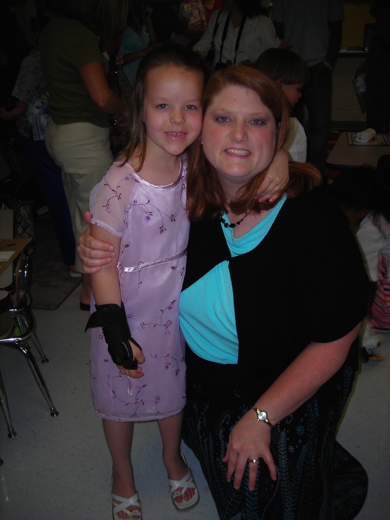 A smiling woman in a black dress stands beside a girl in a pink dress, posing for a photo.