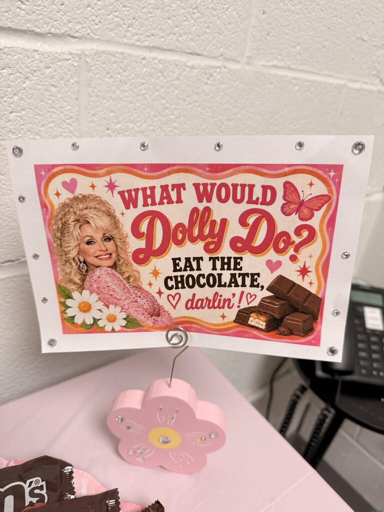 Sign with "What Would Dolly Do?" text and an image of a woman. Below it, a flower-shaped stand holds a chocolate bar.