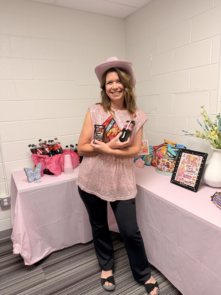 A woman wearing a hat stands next to a table. She holds a bottle and some items.