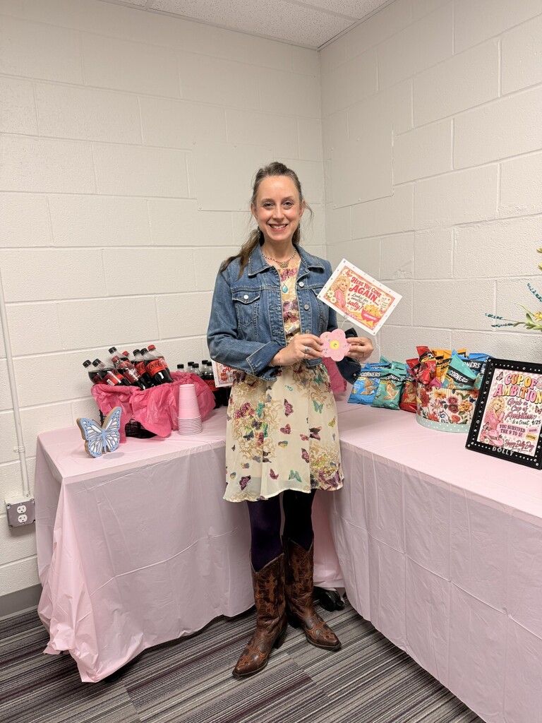 A woman in a floral dress, denim jacket, and boots stands in a room with white walls. She holds a pink card.