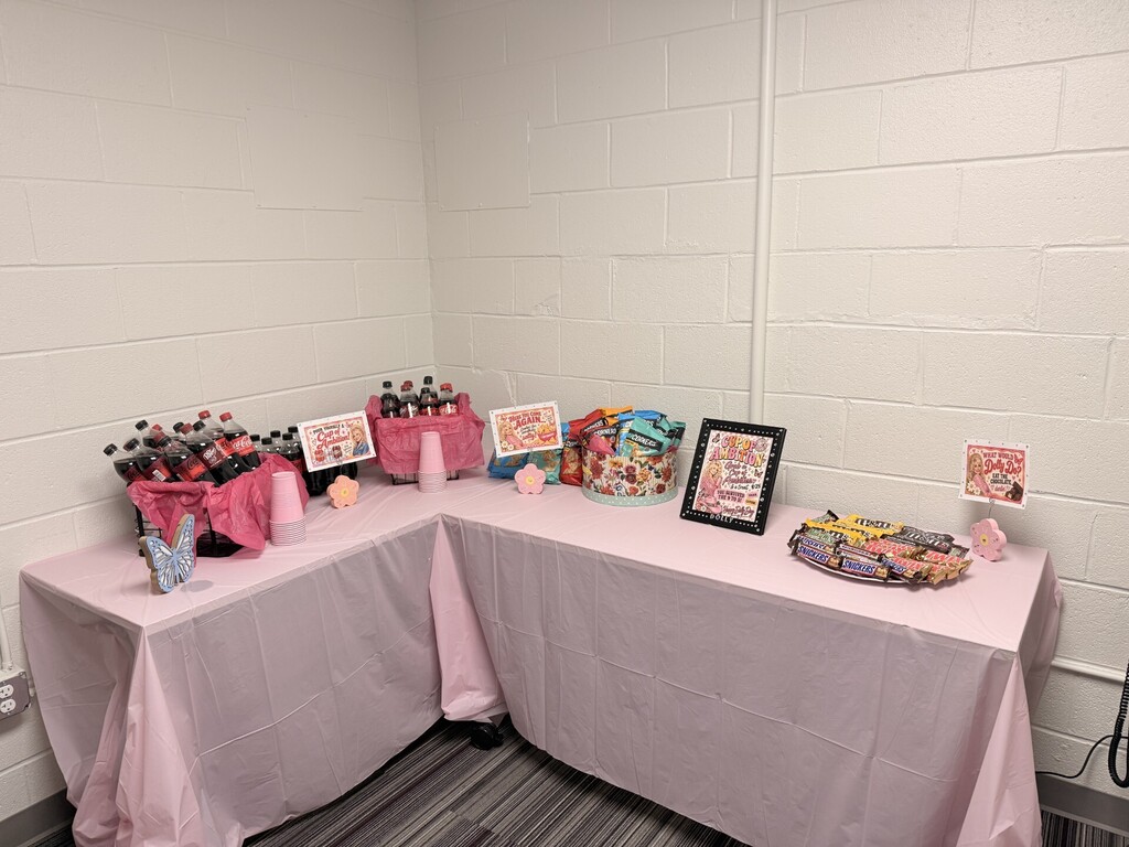 Two tables with pink cloths, holding various items such as baskets, bottles, and books. A white wall is behind the tables.