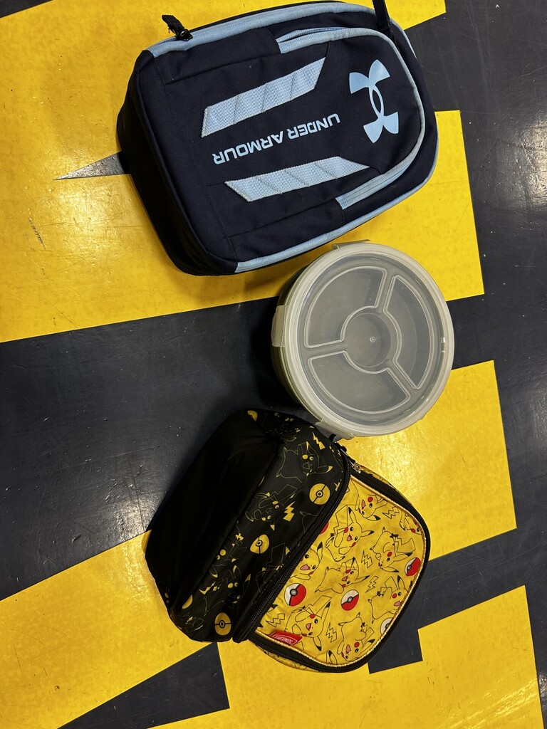 Three bags on a yellow floor with black lines. One is a black lunch bag with the word "Under Armour" in white. Another is yellow with a character design. A grey round container is in between.
