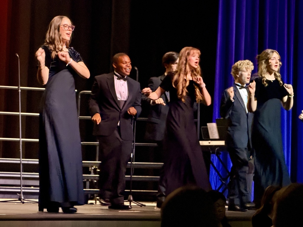 Several people in formal wear stand on stage, facing the audience. Some sing into microphones. The stage has railings and a blue curtain backdrop.