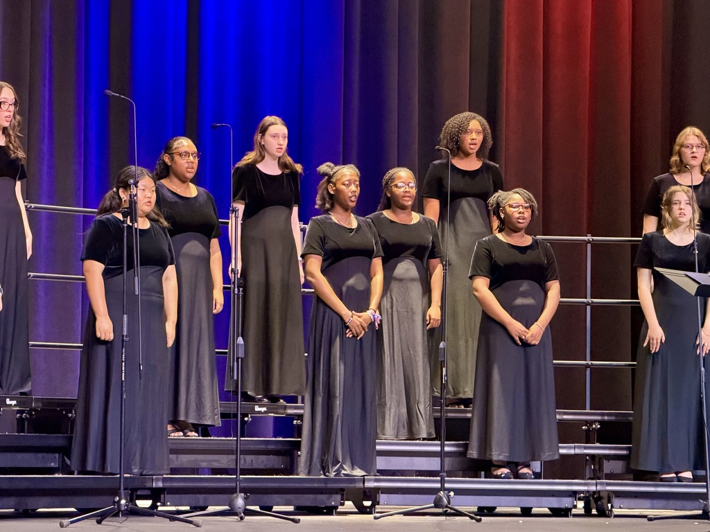 Group of women in matching black dresses standing on stage with microphones in front of a backdrop of blue and red curtains.