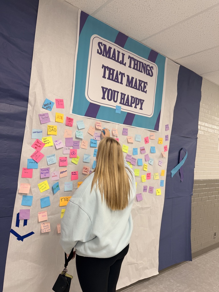 A woman with blonde hair adds a sticky note to a bulletin board with many notes and a banner.