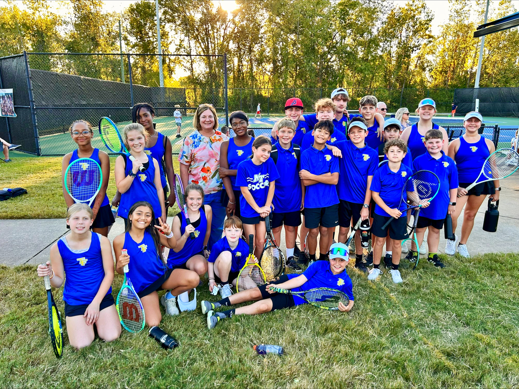 Tennis team in blue uniforms and caps poses for a group photo on a grassy area near a tennis court.