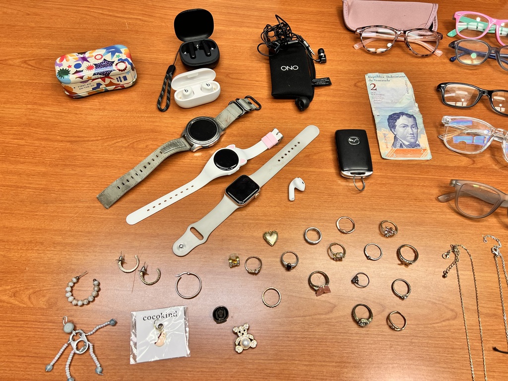 An assortment of jewelry, watches, glasses, a cell phone, earphones, and a wallet on a wooden table.