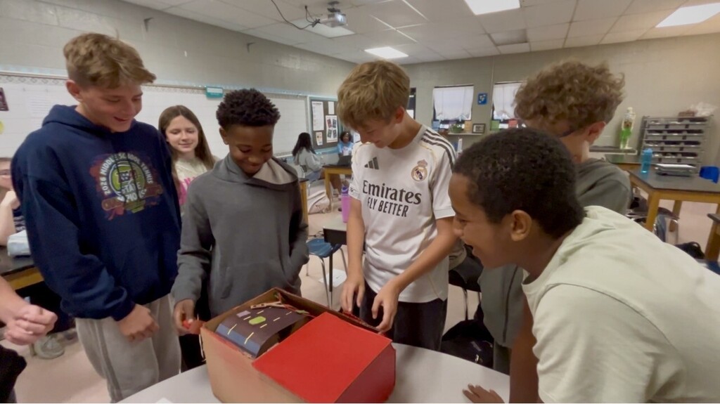 Group of students in a classroom, looking at a box. Two are touching the box.