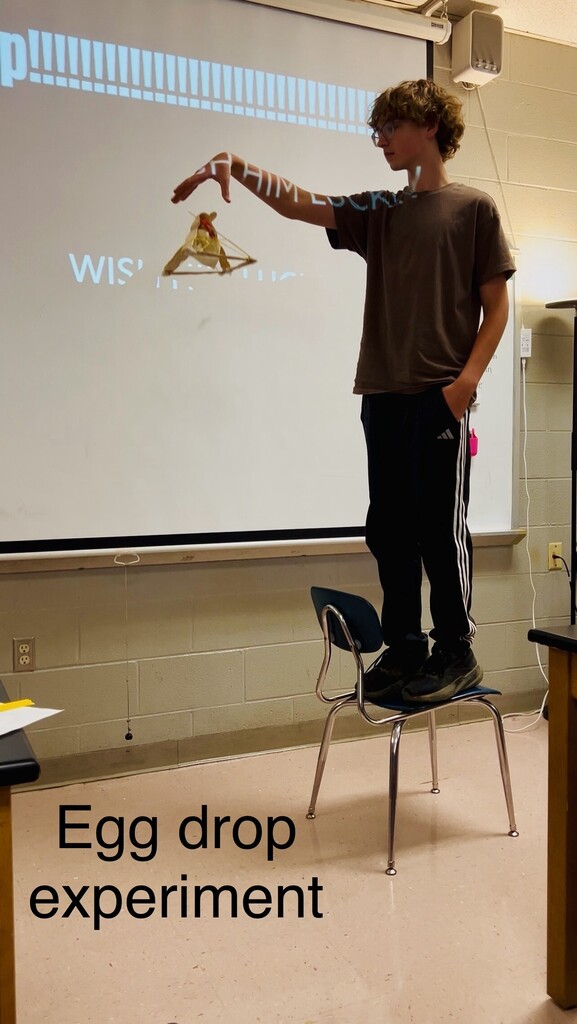 A man standing on a chair in a classroom holding a craft project. Text reads "Egg drop experiment".