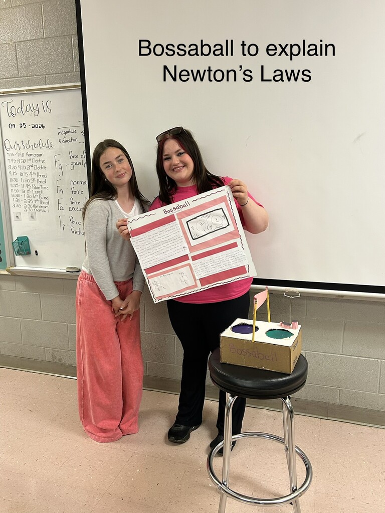 Two women stand in a classroom. One holds a poster and model, the other smiles. A whiteboard is behind them.
