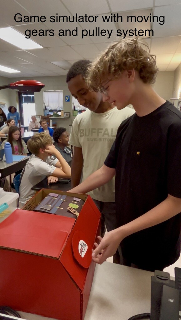 Three boys examine a red box with moving gears and a pulley system. A person sits behind them.