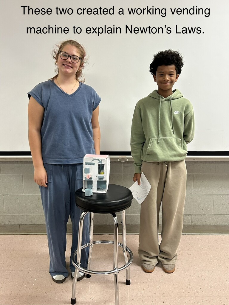 Two students stand in a classroom next to a stool with a small vending machine.