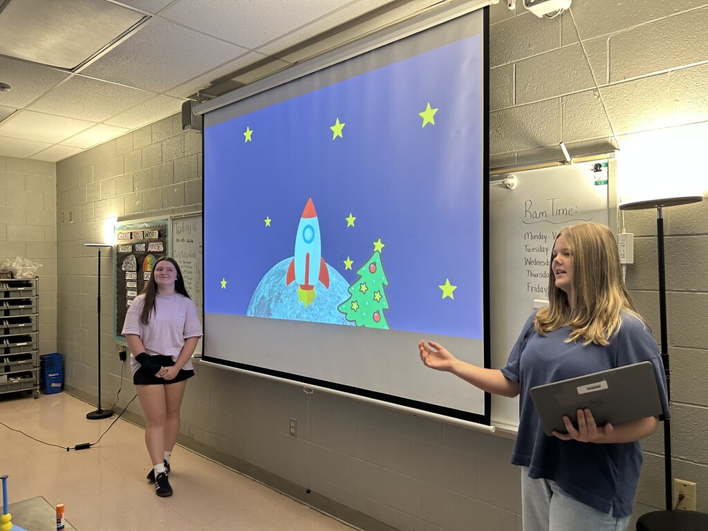 Two women are standing in front of a projector screen displaying a drawing of a rocket. One holds a laptop.
