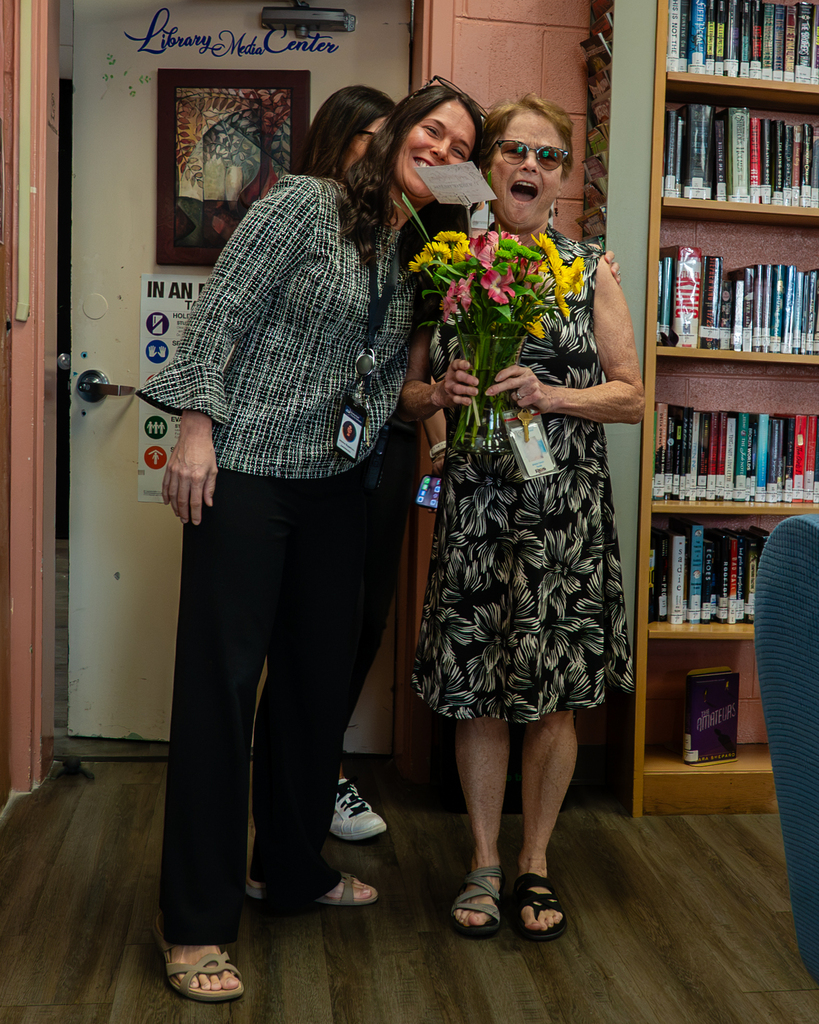 Two women standing in a library hallway with a bouquet of flowers and a shelf of books behind them.