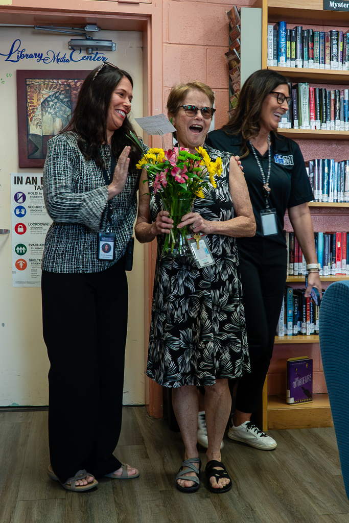 Three women stand in a library. The central woman holds a bouquet of flowers. The woman on the left claps.