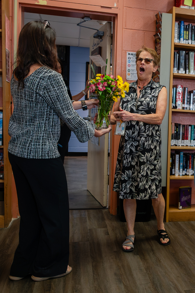 Two women stand inside a room with wooden flooring. One holds a bouquet of flowers and the other holds her mouth in surprise.