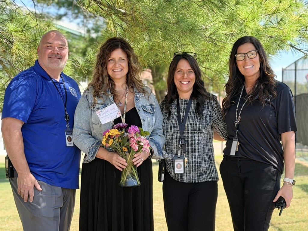Five adults stand in a line under a tree. A woman in a black dress holds a bouquet of flowers.