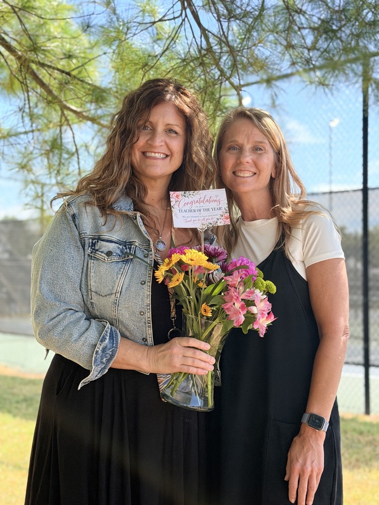 Two women stand under a tree, one holding a bouquet of flowers and the other with a card. Both are smiling.