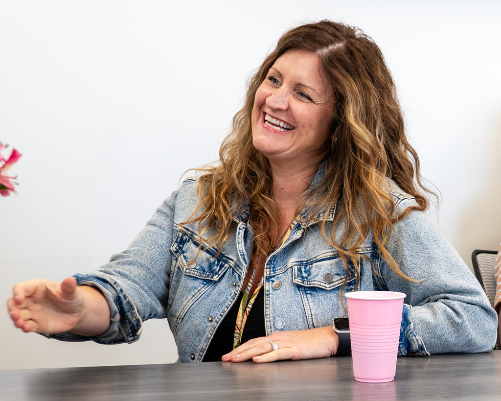 Woman seated at a table with a pink cup. She wears a denim jacket and smiles. Background is plain.