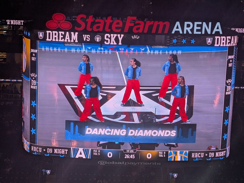 Arena screen displays four women in blue and red costumes performing a dance routine. Logo for State Farm Arena in red.