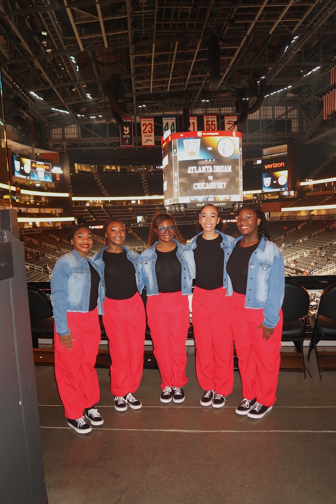 Five women in red pants and black tops stand together in front of an empty stadium, possibly at a basketball event.