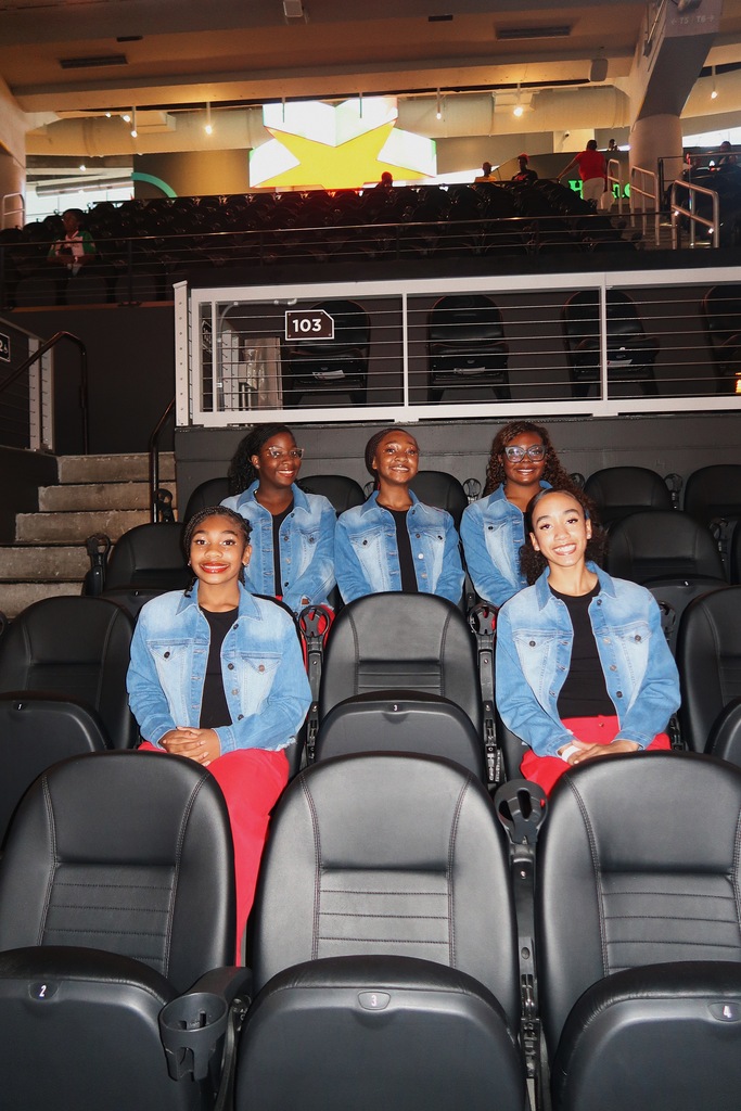 Six women wearing denim jackets and red pants sit together in a theater with empty seats.
