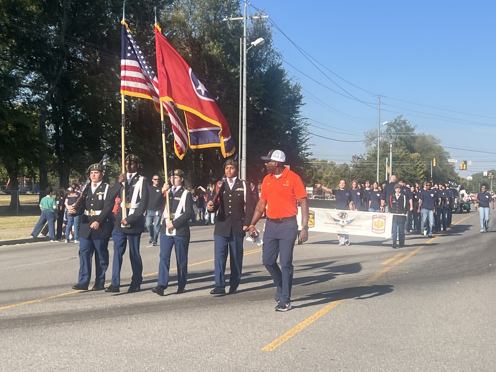 Marching in a parade, people in uniforms carry flags. A man in front wears a hat and orange shirt.
