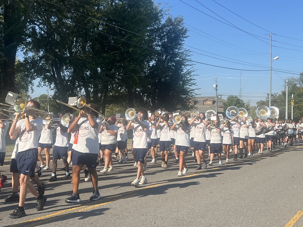 A marching band in white shirts and blue shorts, playing brass instruments, performs on the street.