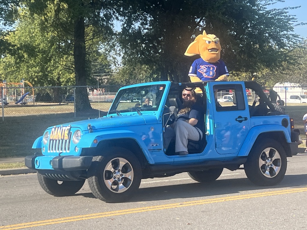 Blue Jeep with mascot on top. Person wearing sunglasses driving. Trees and playground equipment in the background.