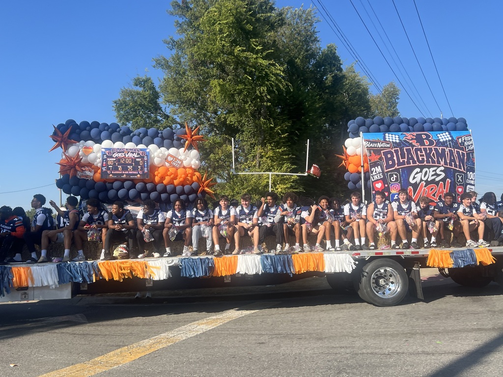 A decorated float travels in the Blackman High School Homecoming Parade. The float features navy, white, and orange balloon displays, football-themed decorations, and large signs reading “Blackman Goes Viral.” Students in football uniforms sit along the sides of the float as it moves down a sunny street lined with trees. Orange, white, and blue fringe decorates the trailer, and a football goalpost prop stands in the center of the float.