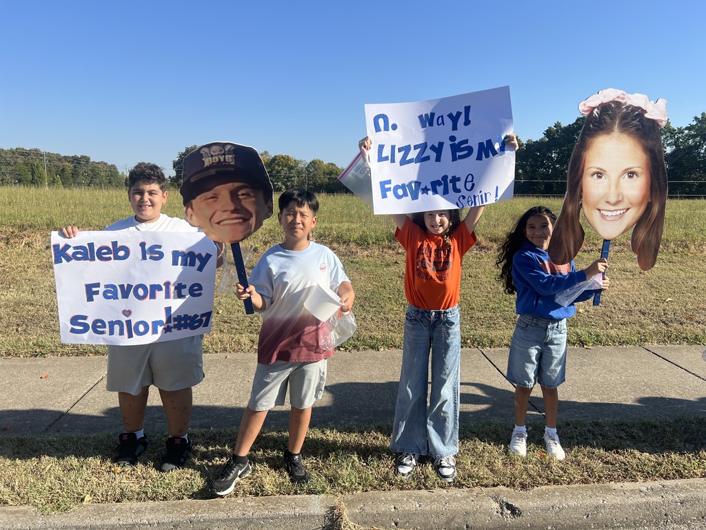 Four children pose with signs on a roadside, holding cutouts of faces. One sign says "Kaleb is my favorite senior."