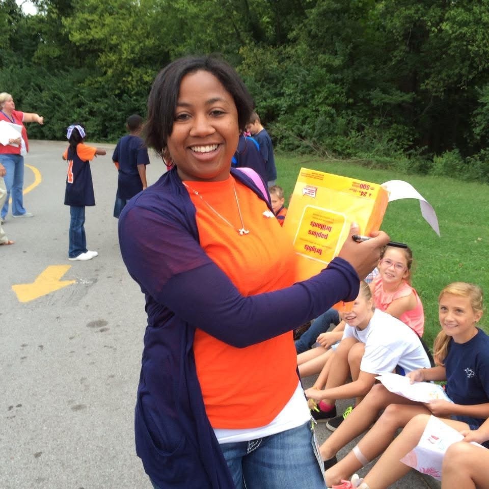 A woman in orange holds a yellow box with a pen and paper outside. Behind her, children sit on grass.