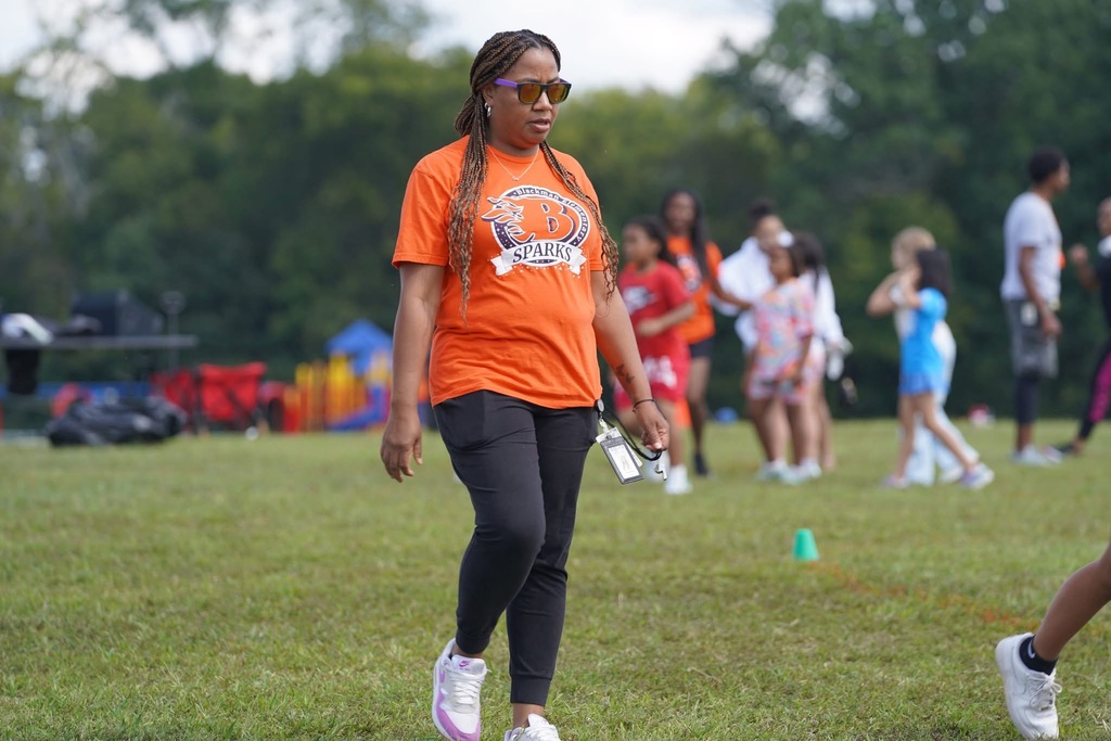 Woman in orange shirt and black pants walks across grass. Children in background. Green grass, trees, picnic table.